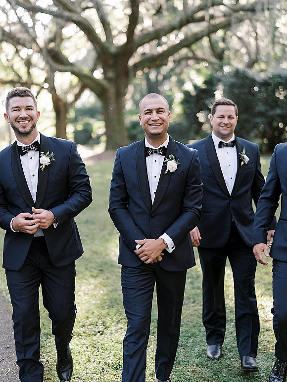 Groomsmen portrait of men in navy tuxedos with black bow ties and white boutonnieres under a large oak tree in dappled sunlight