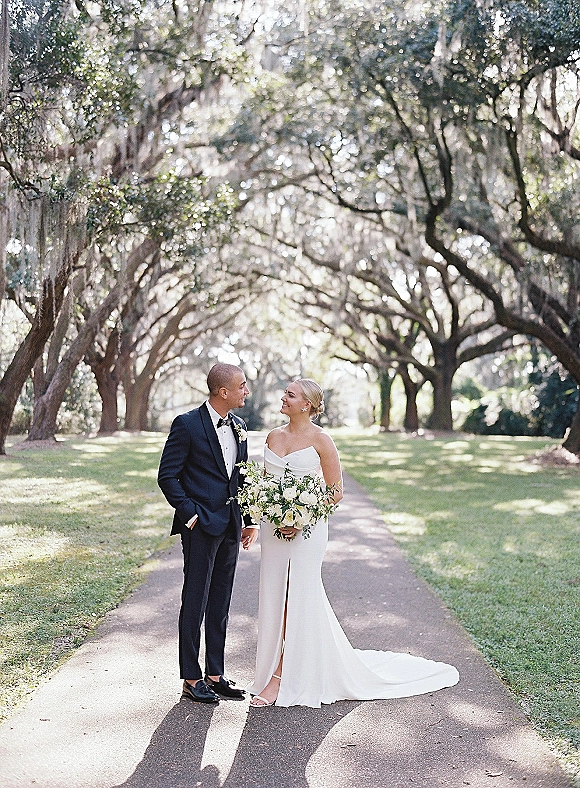 Couple portrait of bride and groom holding hands, her white and green bouquet and strapless gown on a tree-lined path in dappled sunlight
