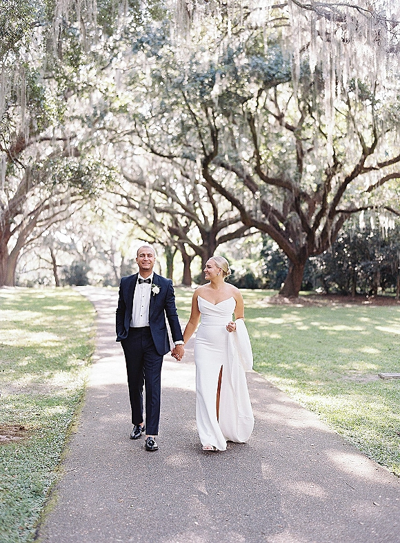 Couple portrait of bride and groom walking hand in hand, bride in strapless slit gown looking at him under mossy trees on a path