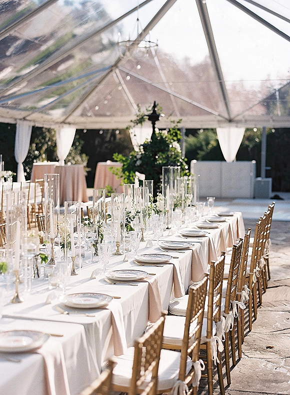 Reception tablescape with a long banquet table wedding setup, white linens, gold chiavari chairs, taper candles, and greenery under a clear tent