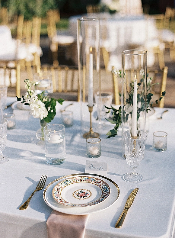 Reception tablescape with blush napkin, floral china plate, gold flatware, crystal goblet, and candlelight on an outdoor wedding table setting