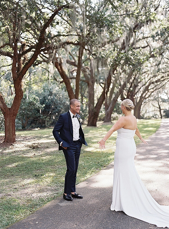 Wedding first look as bride in a strapless dress approaches groom in tuxedo on a tree-lined path with hanging moss in dappled sun