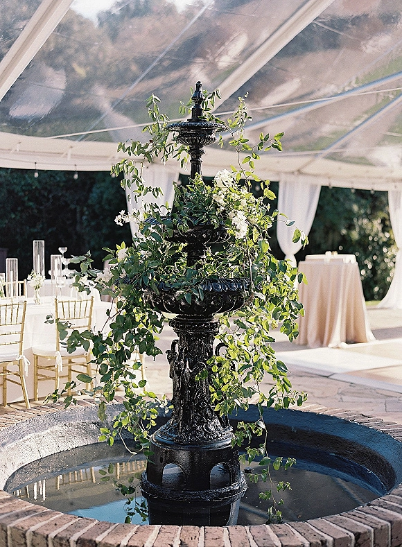 Wedding fountain decor with a tiered black fountain wrapped in greenery and white flowers, set under a clear tent on a sunny patio