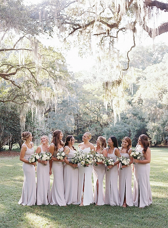 Bridesmaid group photo of women in mismatched neutral dresses holding white rose and greenery bouquets under oak trees with Spanish moss