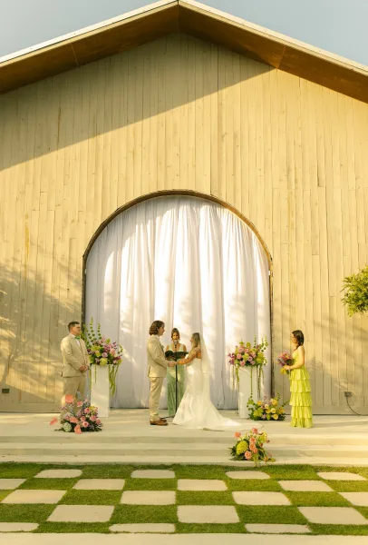 Wedding ceremony with couple exchanging vows under a white draped backdrop, surrounded by floral pedestal stands before a barn facade