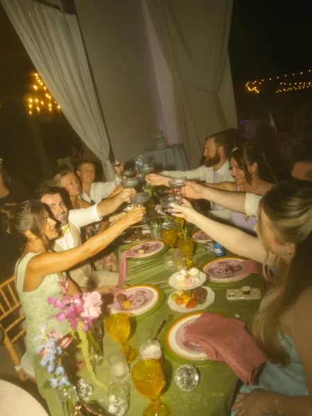 Wedding reception toast with guests raising champagne coupes amid candlelit tablescape, floral centerpiece, and string lights in a draped tent at night