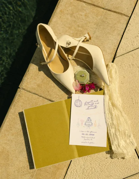 Invitation suite wedding invitation flat lay with cream bridal heels, engagement ring in green velvet box, and petals on stone steps by water