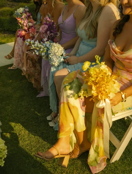 Bridesmaids portrait of women sitting on a bench holding bouquets, pastel mismatched dresses with ribbon wraps on a grass lawn near ceremony seats