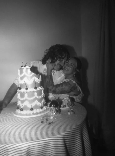 Wedding cake cutting as bride and groom slice a cherry-topped white tiered cake at an indoor reception table against a plain wall
