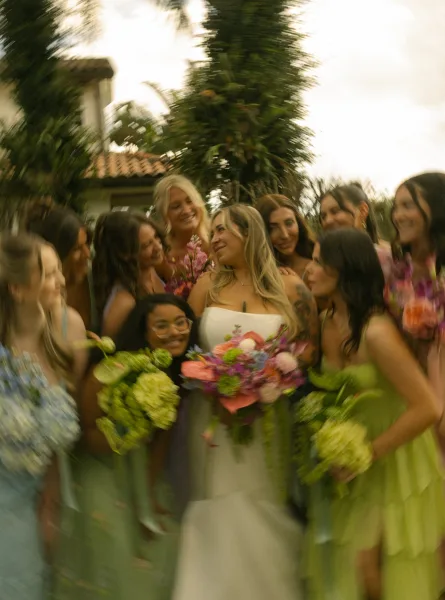 Bride with bridesmaids in mismatched dresses holding colorful bouquets and wine glasses, posing in a palm-lined garden by a stucco building