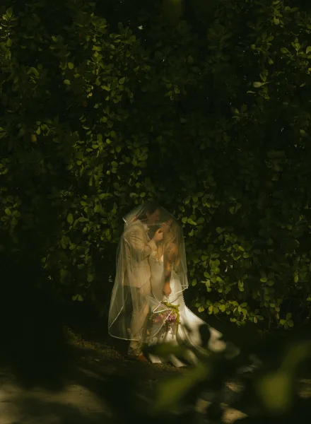 Wedding couple portrait of bride and groom under veil, touching foreheads as she holds a greenery bouquet before a lush hedge in dappled sunlight