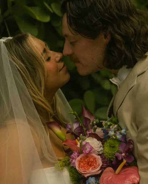 Wedding couple portrait with bride and groom nose to nose under a bridal veil, holding a colorful bouquet against lush green foliage