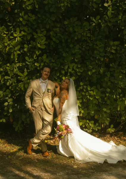 Couple portrait of bride and groom posing as she leans on him, holding a bouquet, in dappled sunlight before a lush greenery wall