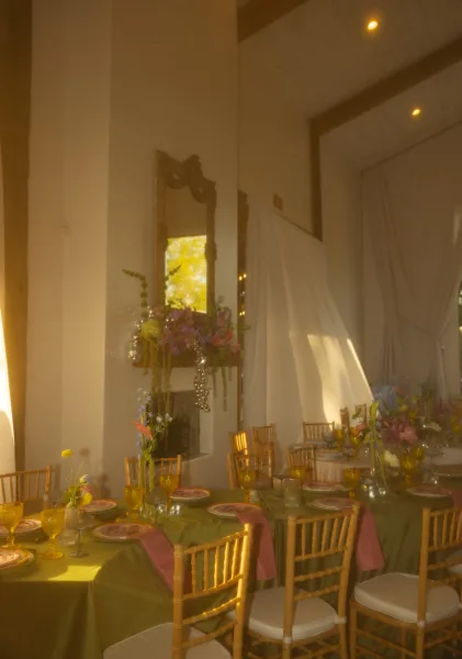 Reception tablescape with round reception tables in green cloth, colorful centerpieces, amber goblets and pink napkins in a tall white hall