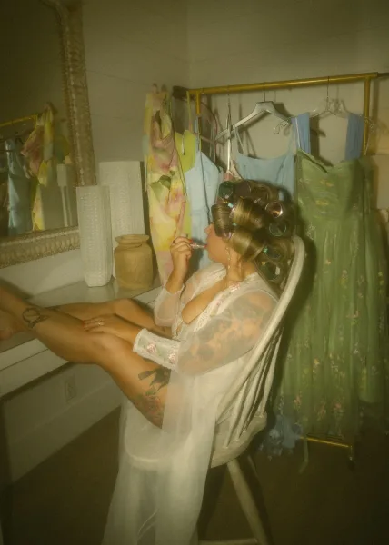 Bridal getting ready as the bride puts on lipstick in hair rollers, wearing a robe beside a bathroom vanity mirror with dresses on a rack