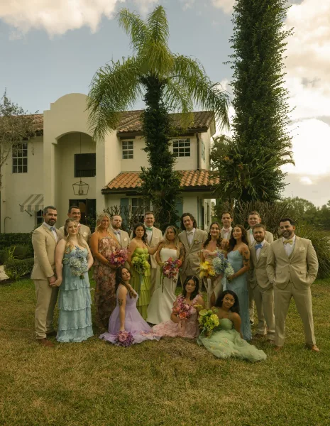 Wedding party portrait with bride, bridesmaids and groomsmen posing on a lawn, holding bright bouquets beside a stucco villa and palm trees