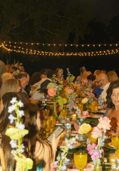 Reception tablescape at an outdoor wedding reception on a long banquet table, candlelit with string lights, florals, and gold chiavari chairs under trees