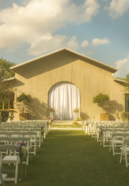 Ceremony setup with white folding chairs lining a lawn aisle, pastel florals and white draped backdrop with chapel facade under blue sky