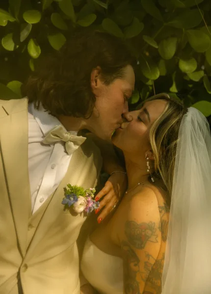 Wedding kiss close up as the bride in a veil and pearl drop earrings embraces the groom in a bow tie, with green foliage behind