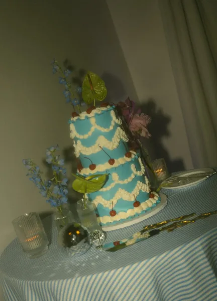 Wedding cake with blue icing and white piped frosting, topped with cherries and tropical leaves, on a striped tablecloth with votives