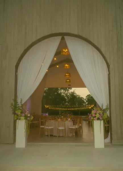 Reception entrance decor with draped barn entrance, white curtains and pastel floral pedestals glowing under bistro lights at dusk in an open-air pavilion