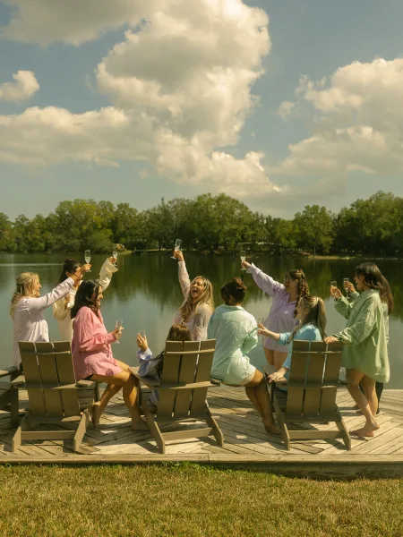 Bridesmaids toast with champagne flutes in oversized button-down shirts, laughing on Adirondack chairs by a lake dock under clouds