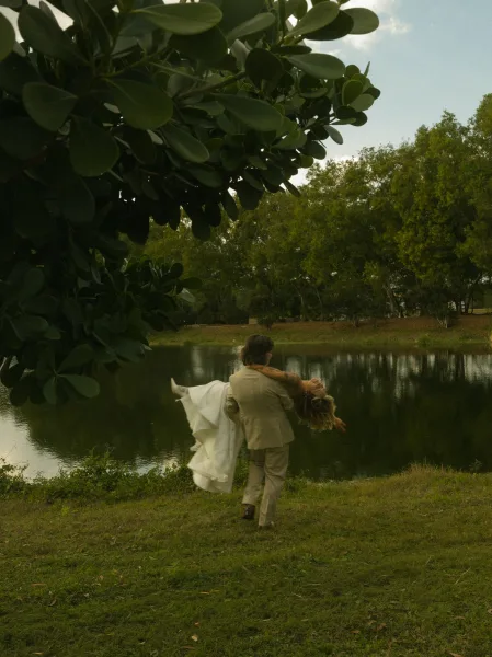 Couple portrait of groom carrying bride in a wedding dress with white heels and tattoos, walking by a lakeside under tree branches