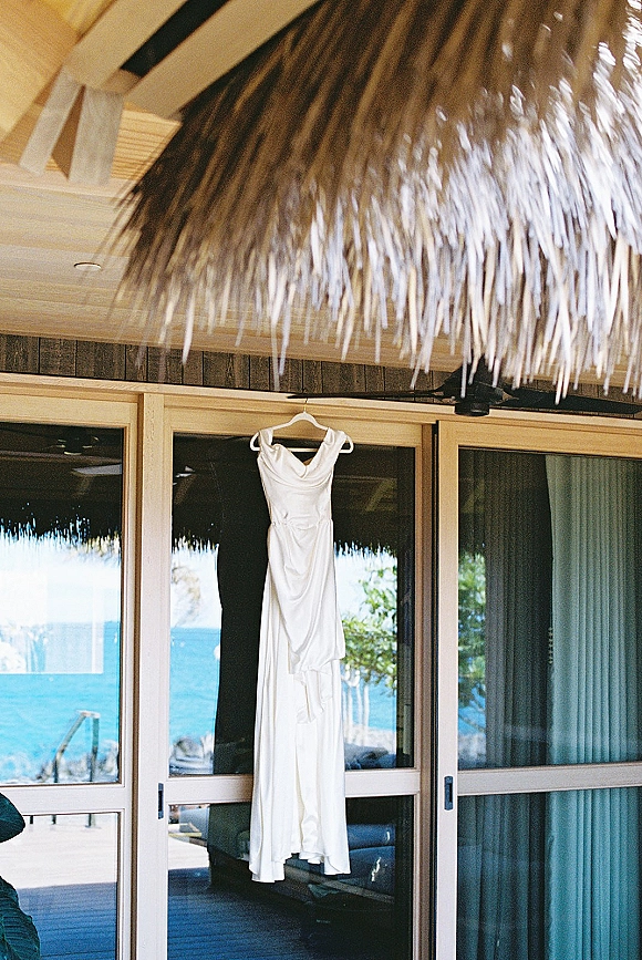 Wedding dress on hanger in soft natural light by sliding glass doors, with ocean view beyond a wooden deck and thatched roof