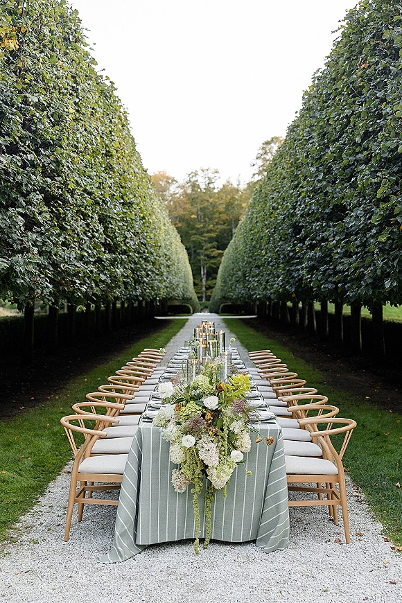 Outdoor reception table with a long banquet table setup, striped linens, white-flower garland, and taper candles in a tree-lined garden alley
