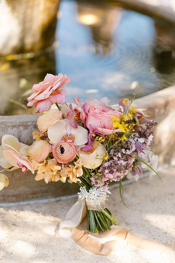 Bridal bouquet of pink roses, orchids, ranunculus and lilac blooms with greenery and a jeweled brooch, resting by a stone fountain edge
