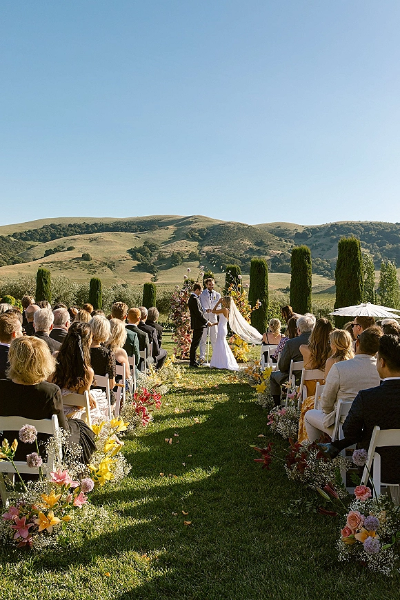 Wedding ceremony as bride and groom exchange vows under a floral arch, with guests seated along a petal-strewn aisle in a vineyard setting