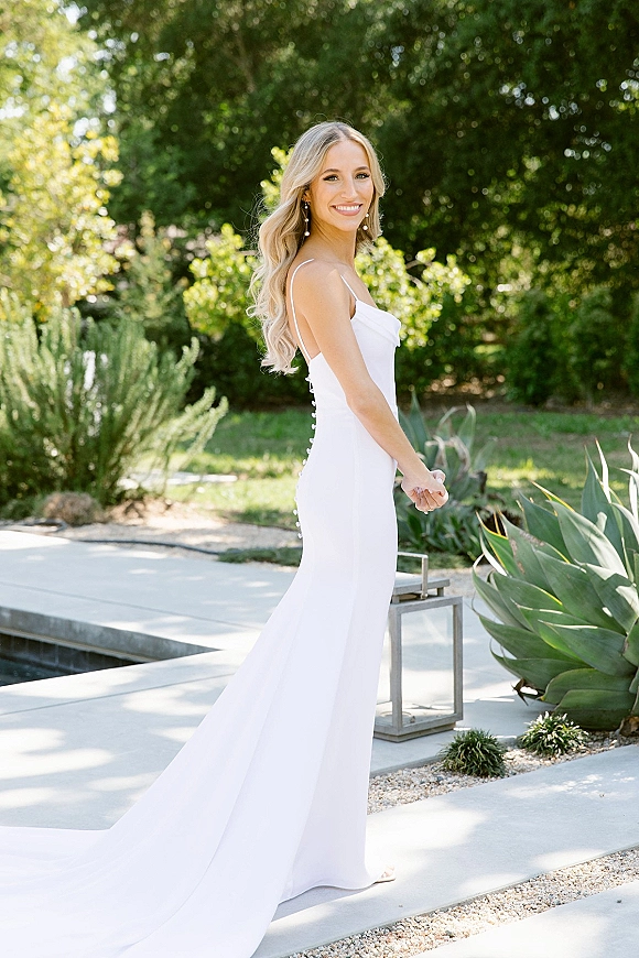 Bridal portrait of a smiling bride looking back in a low-back spaghetti strap gown with pearl drop earrings on a garden path