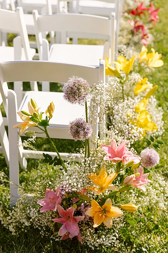 Ceremony aisle decor with outdoor ceremony chairs in white folding chair rows, framed by low lilies and baby's breath on a grass lawn