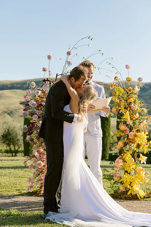 Ceremony kiss at an outdoor wedding ceremony beneath a floral arch, bride in strapless gown and long veil, hills and blue sky behind