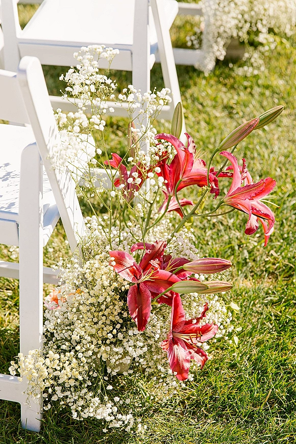 Ceremony aisle flowers with baby’s breath and pink lilies beside white folding chairs on a grass lawn at an outdoor ceremony space