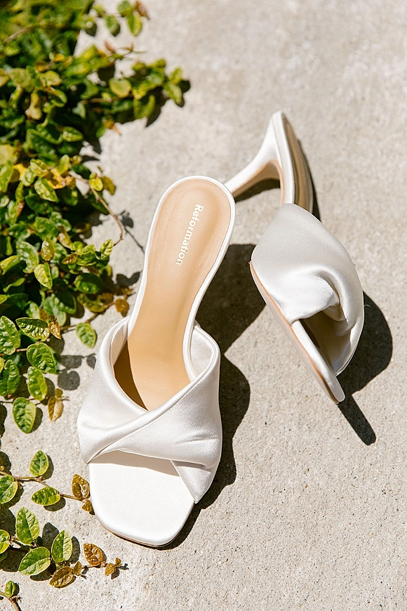 Bridal shoes, white satin wedding heels with twisted strap detail and stiletto heel on concrete with green vines and sunlit shadows