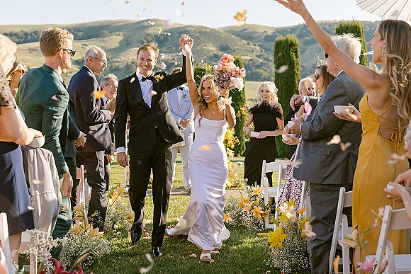 Wedding recessional as newlyweds walking down aisle, bride lifts bouquet overhead beside groom in tuxedo, petals fly on mountain lawn with cypress trees