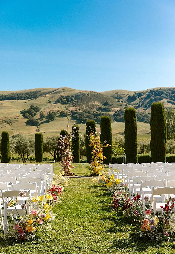 Outdoor ceremony setup with a wedding ceremony aisle lined in lilies and baby's breath, white chairs on a vineyard lawn with hills and cypress trees.