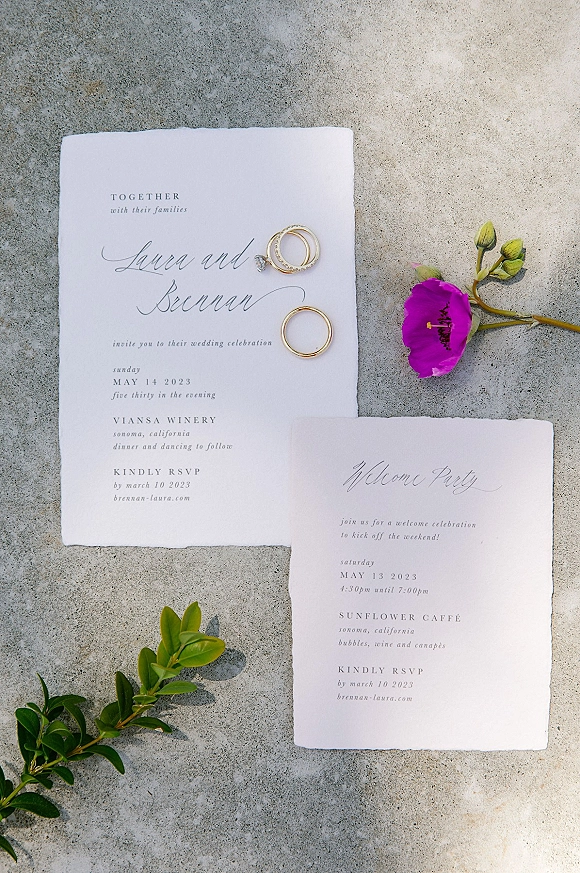 Invitation suite wedding invitation flat lay with deckled edge paper, calligraphy text, wedding rings, and a pink bud on concrete surface