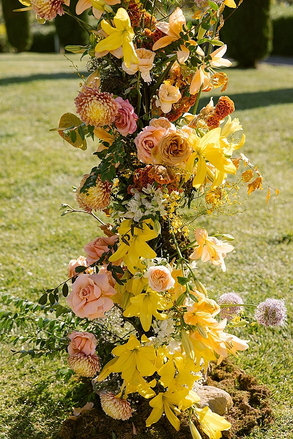 Wedding floral arrangement with yellow lilies and peach roses nestled in greenery on rocks along a sunlit lawn with hedges