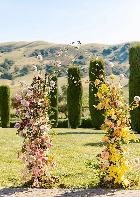 Ceremony floral pillars with roses and orchids framing an outdoor aisle entrance, set against cypress trees and a mountain view under blue sky