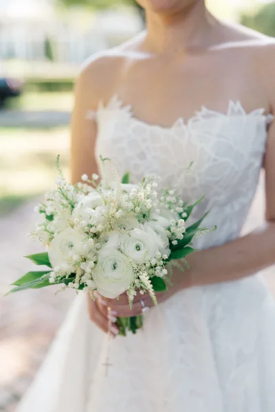 Bridal bouquet of white flowers and lily of the valley with greenery, held against a strapless lace dress on an outdoor walkway