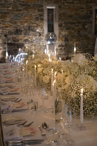 Reception tablescape styled as a candlelit wedding table with white roses, set against a stone wall beneath a stained glass window.