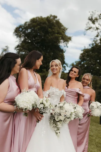 Bridal party portrait of a bride with bridesmaids, holding bouquets on a garden lawn under a cloudy sky.