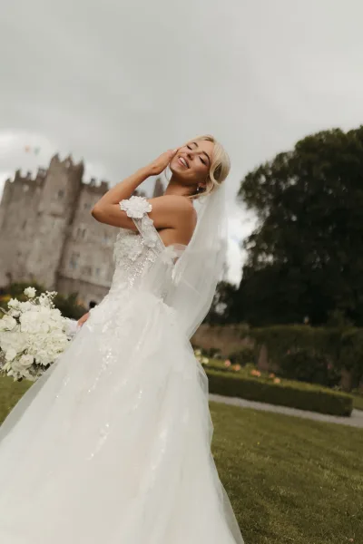 Bridal portrait of a bride with bouquet, posed in a castle garden with clipped hedges and a broad lawn beneath a softly cloudy sky.