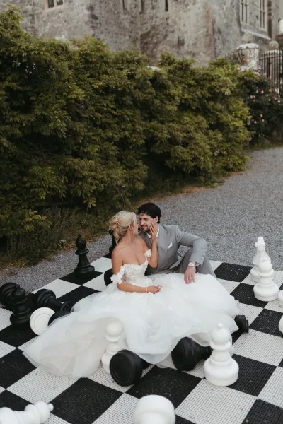 Couple portrait of a bride and groom sitting close beside giant chess pieces, she touches his face in a candid moment.