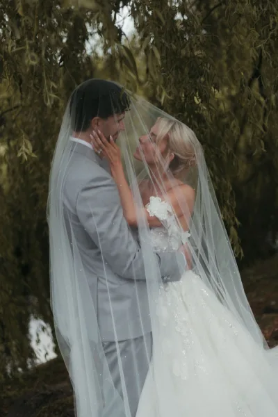Couple portrait, an under the veil photo as the bride and groom embrace beneath willow branches by the water.