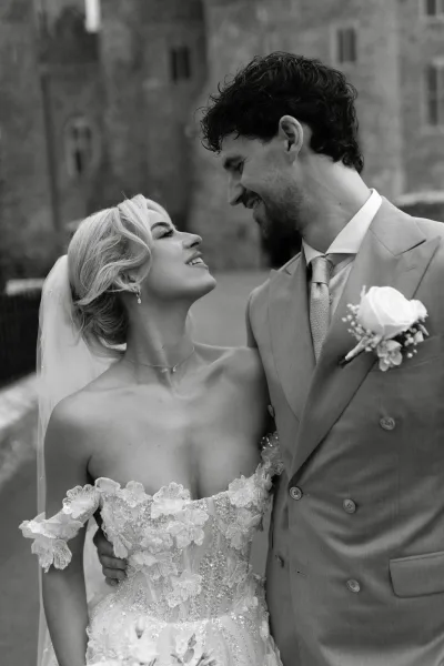 Couple portrait as the bride and groom embrace, her veil catching light as they smile beside a stone building and fence along a quiet walkway.