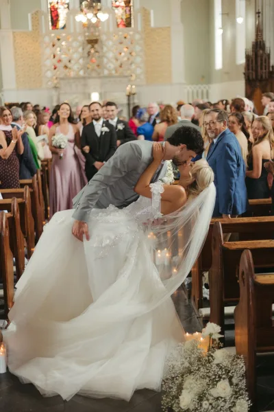 Wedding kiss portrait of a ceremony recessional kiss as the groom dips the bride beneath stained glass, her veil trailing down the aisle.