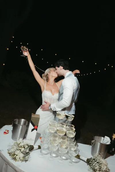 Wedding kiss portrait of the couple sharing a champagne tower kiss in a garden at night, trees silhouetted against the dark sky.